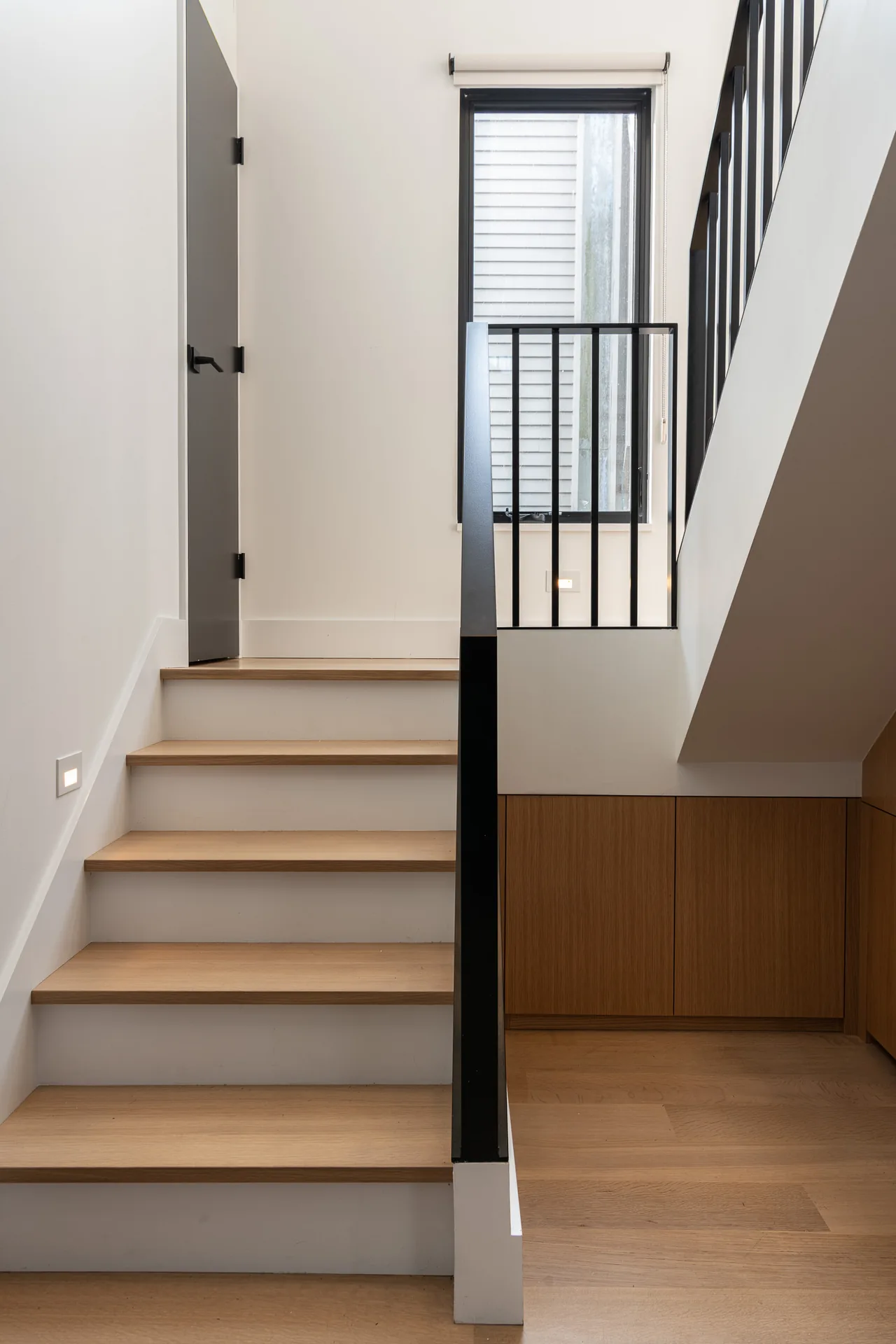 Staircase with light wood treads and black metal railing beside a tall window with black trim.