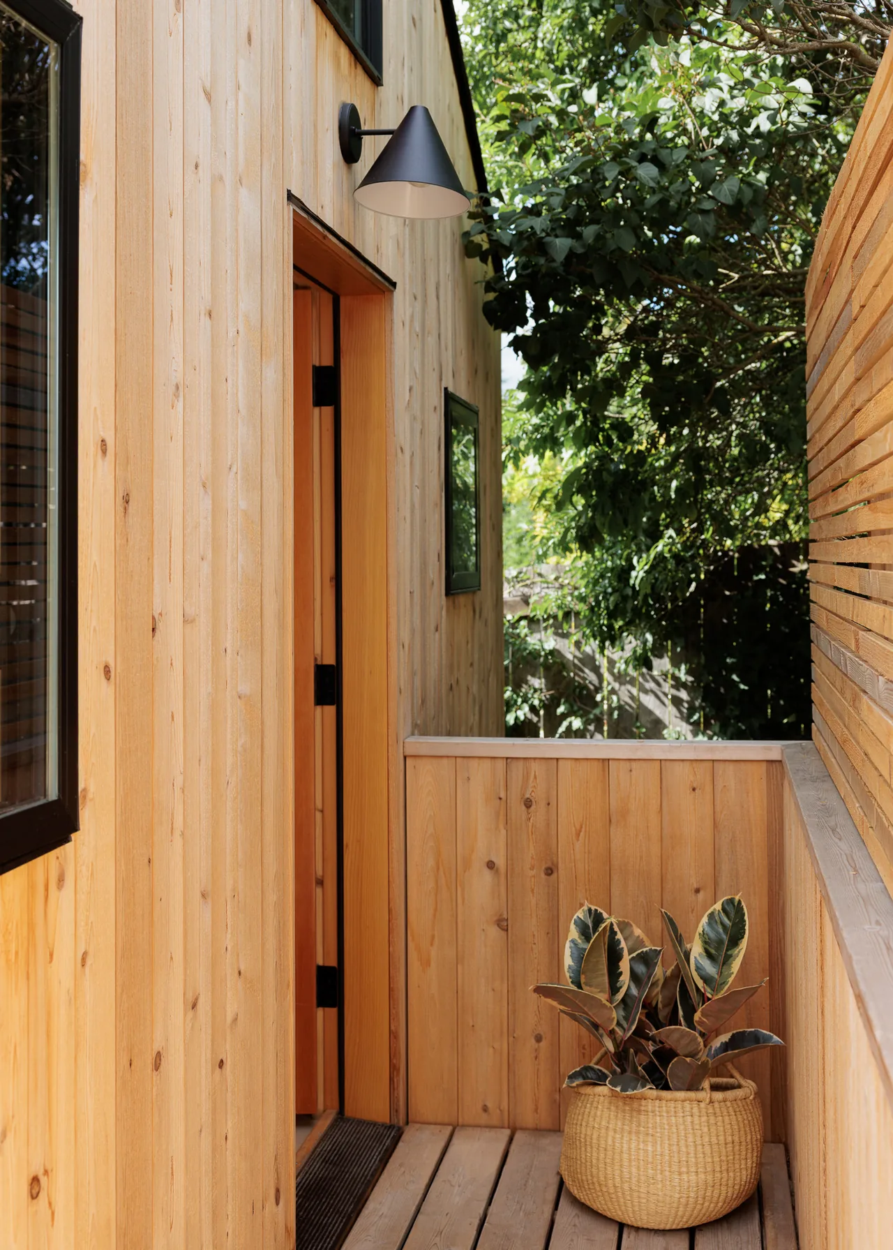 Outdoor walkway space clad in natural wood siding with a black wall sconce and a potted plant near the entrance.