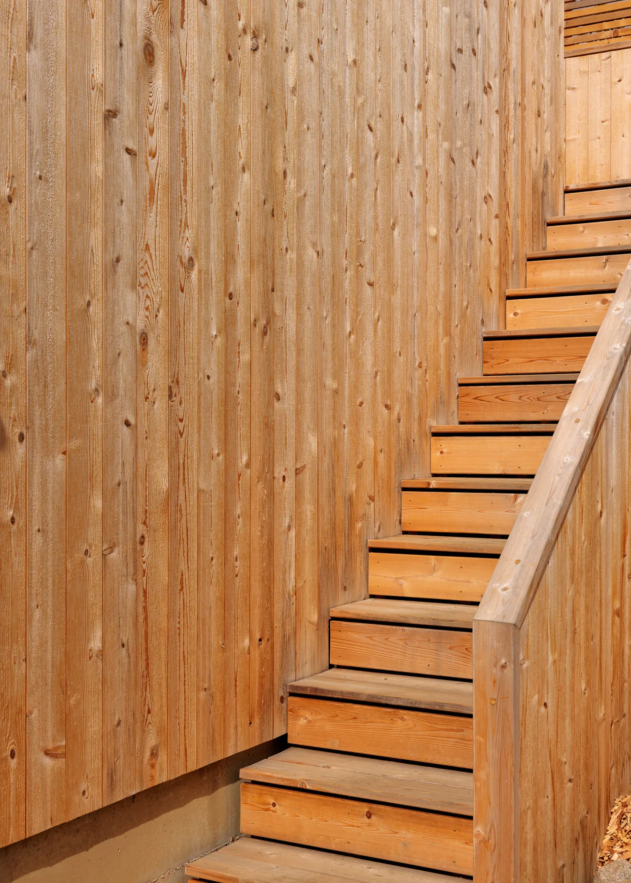 Outdoor wooden stairs with a matching wood railing rising alongside vertical wood siding.