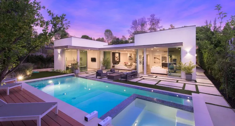 Modern single-story house with large glass sliding doors opening to a backyard patio and swimming pool at dusk.