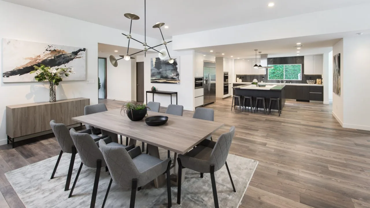 Dining room with gray upholstered chairs and modern chandelier adjacent to an open kitchen with a large island and stainless steel fridge.