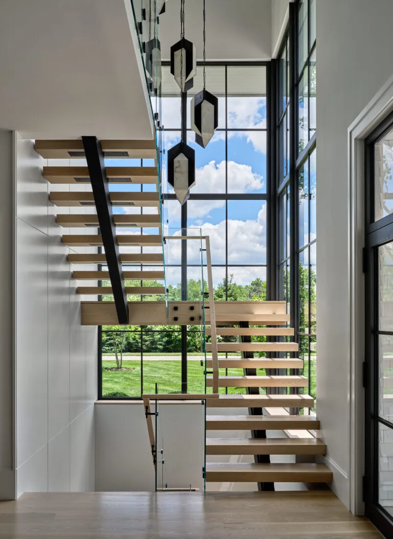 Floating wood stair with glass railing beside expansive black grid windows and pendant lights in a two-story space.
