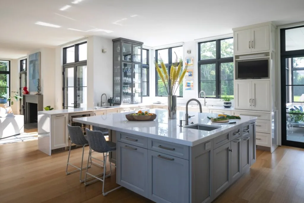 Contemporary kitchen featuring a large island with sink, black-framed tall windows, and light wood flooring.