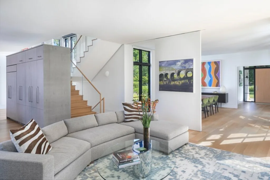 Open living room with curved gray sectional, glass table, light wood flooring, and adjoining dining area with green chairs.