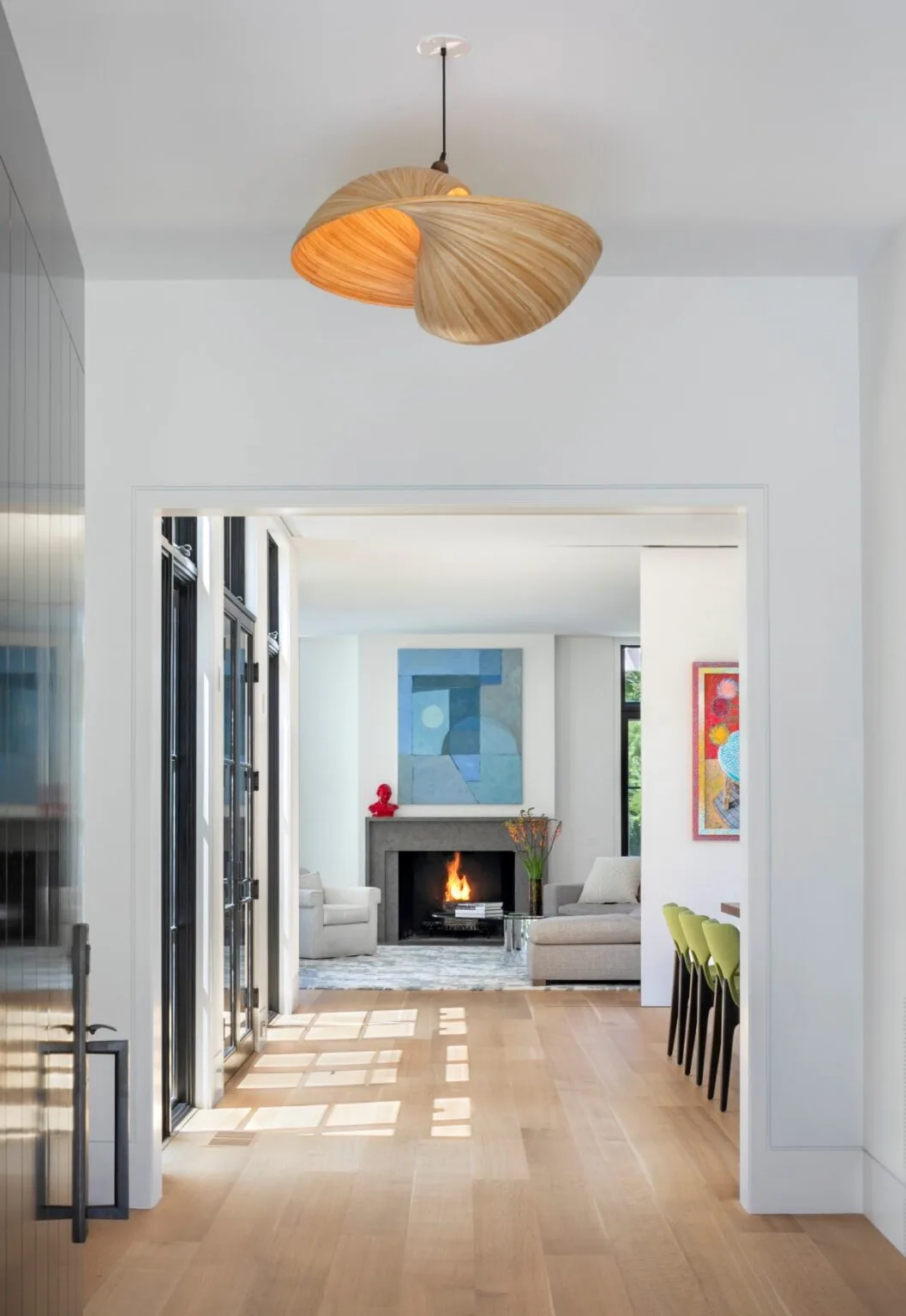 Hallway view into a modern living room featuring a lit fireplace, contemporary art, and light wood flooring.