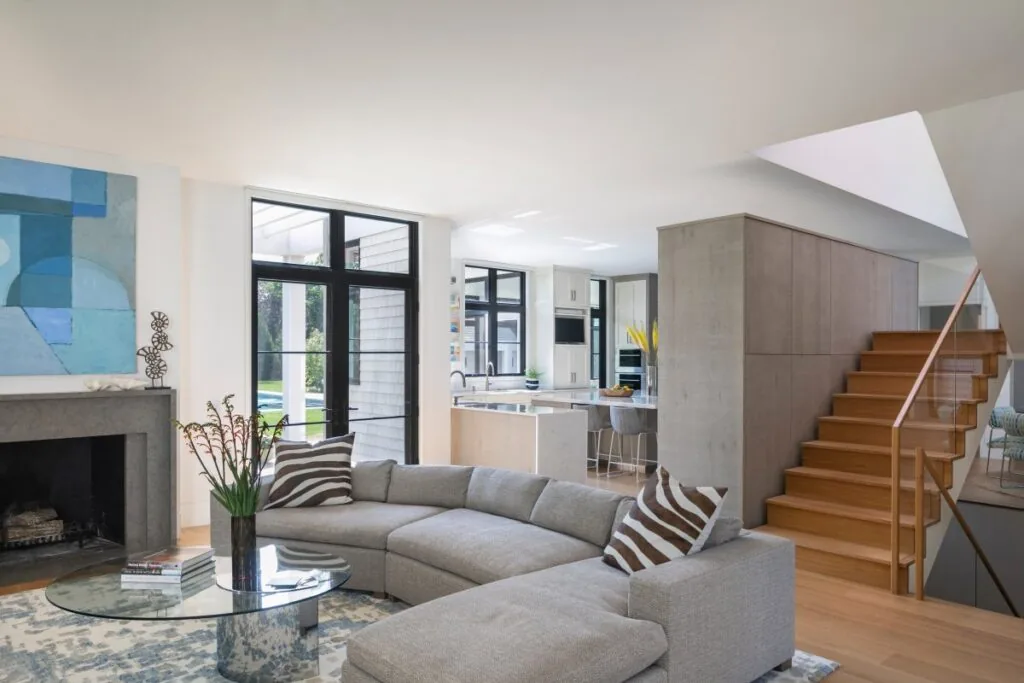 Open living room with sectional sofa, glass coffee table, fireplace, and view into kitchen with bar stools and wooden stairs.