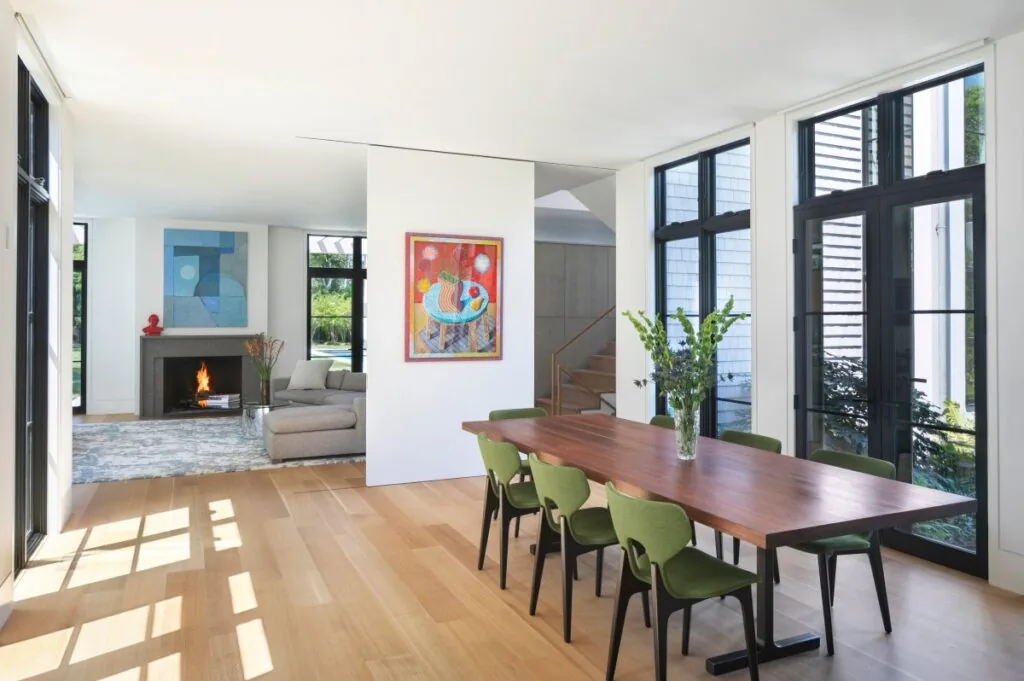 Dining room with green chairs and a wood table next to tall black window frames, adjacent to a living room featuring a fireplace and artwork.