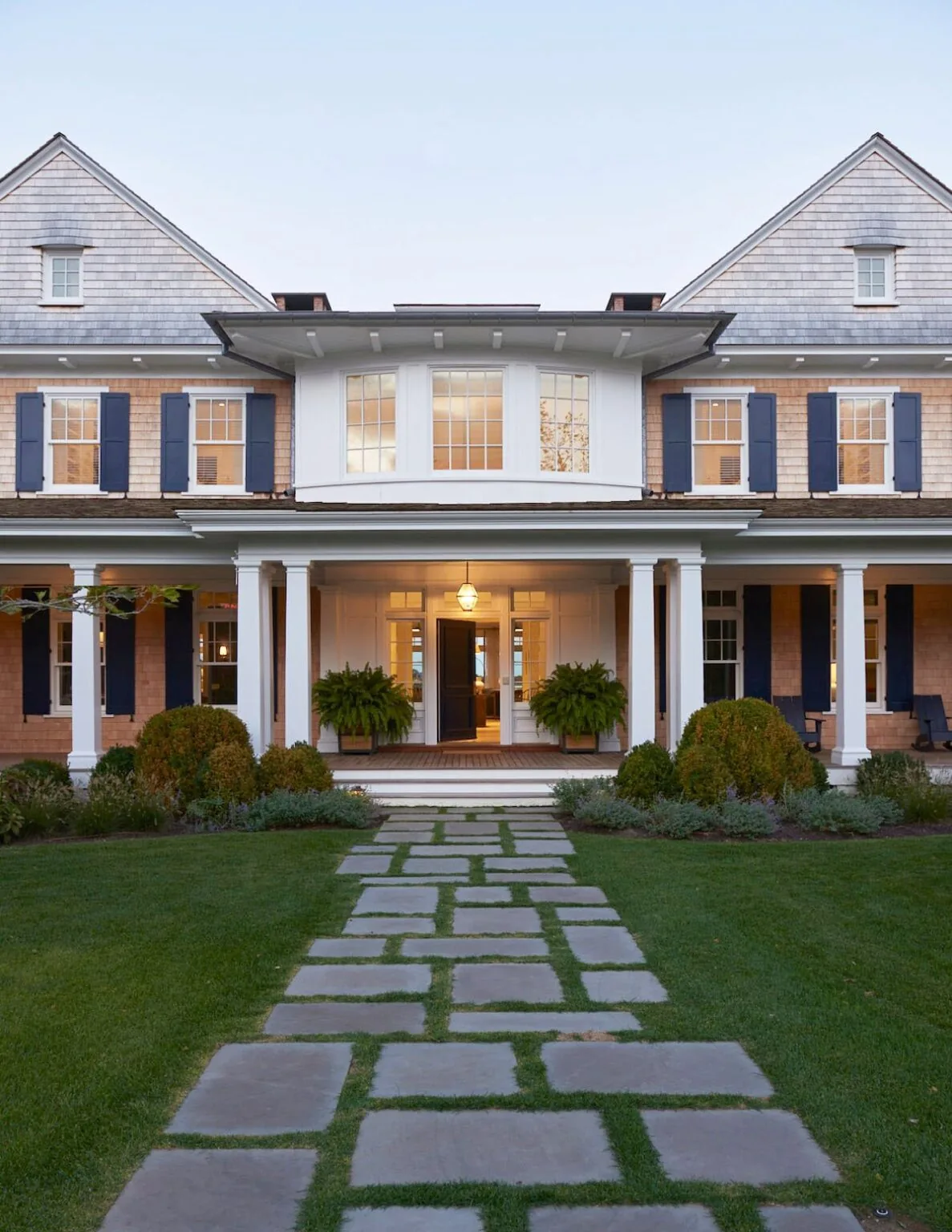 Traditional two-story house with shingle siding, multiple double-hung windows with shutters, and a front porch with columns and a hanging lantern.