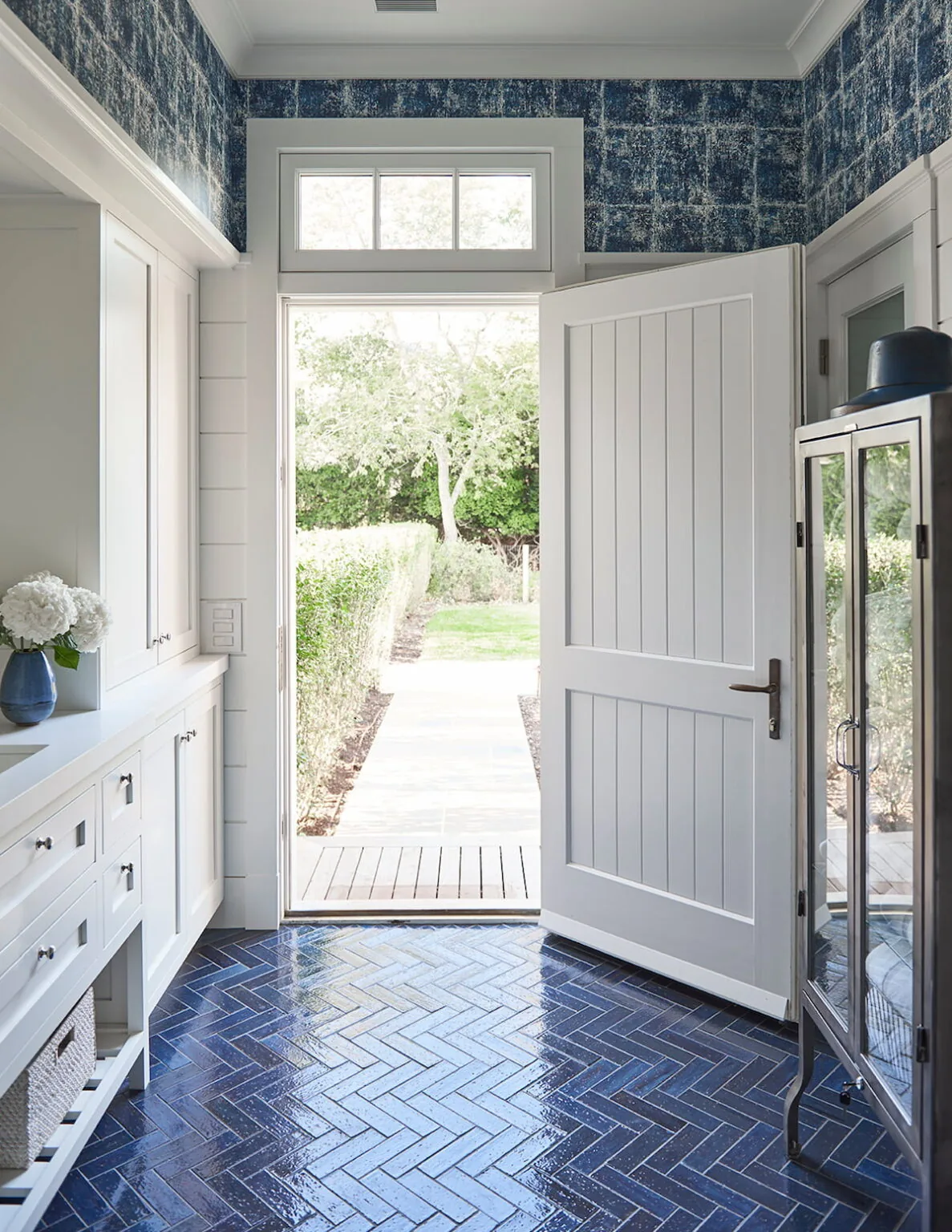 Entryway with navy blue floor tiles, beadboard walls, and white cabinetry opens to a garden path.