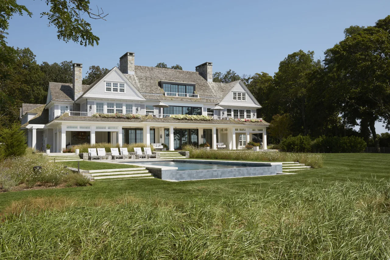 North Fork on the Water features a traditional white house with stone roofing, multiple chimneys, and a formal rectangular pool in a spacious yard.