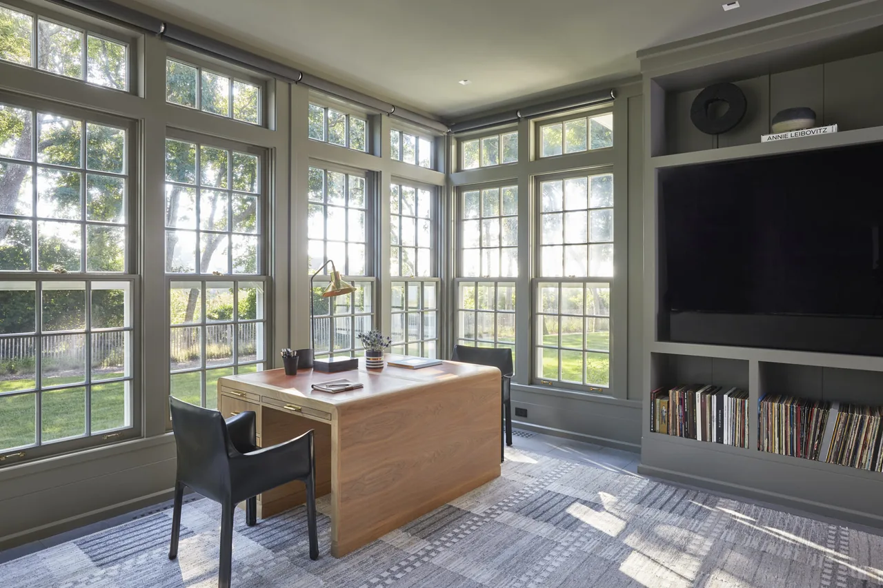 Home office corner with wood desk, black chairs, large multi-pane windows, and built-in shelving with books and a TV.