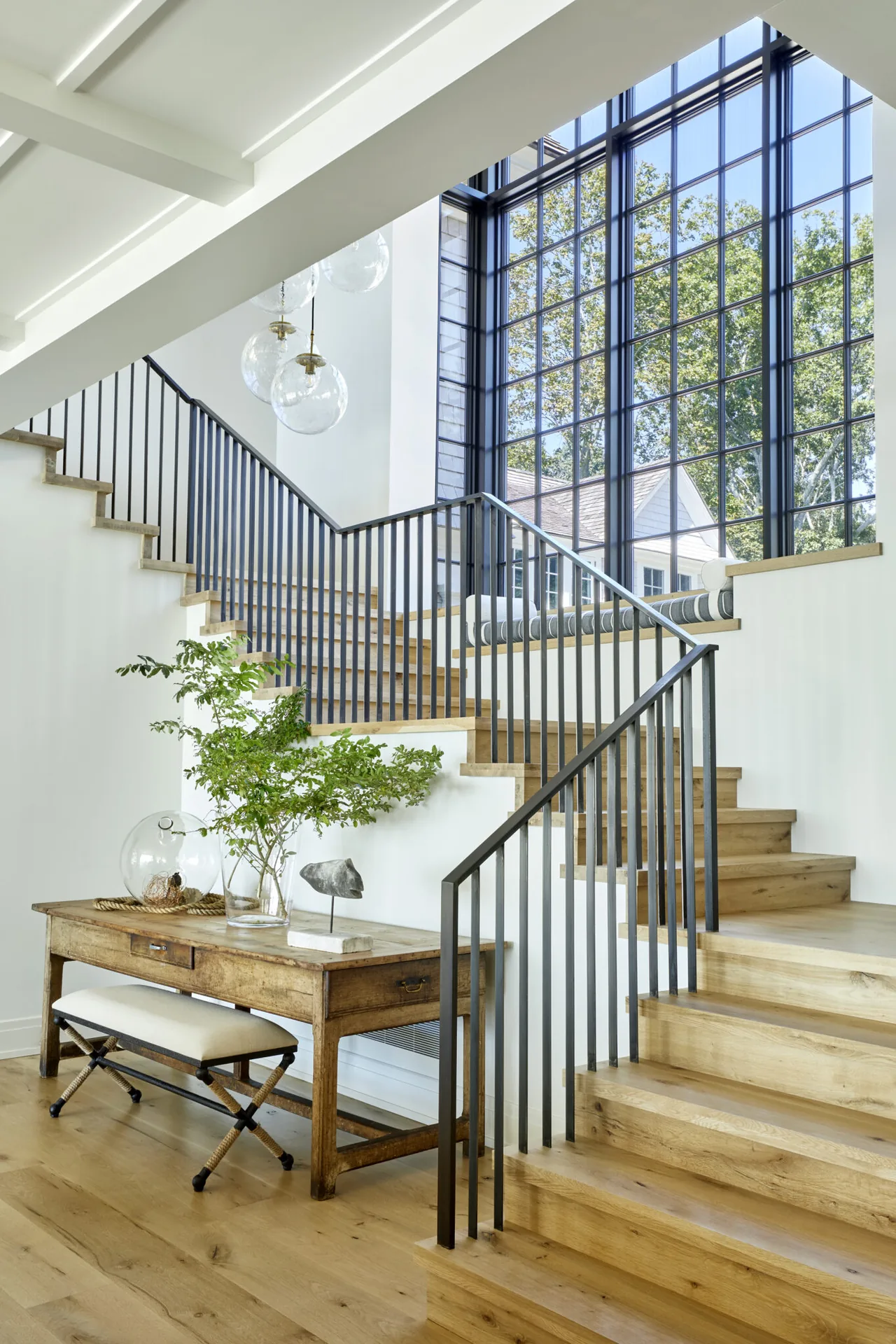 Modern staircase with wood treads and black metal railing next to large grid windows overlooking trees.
