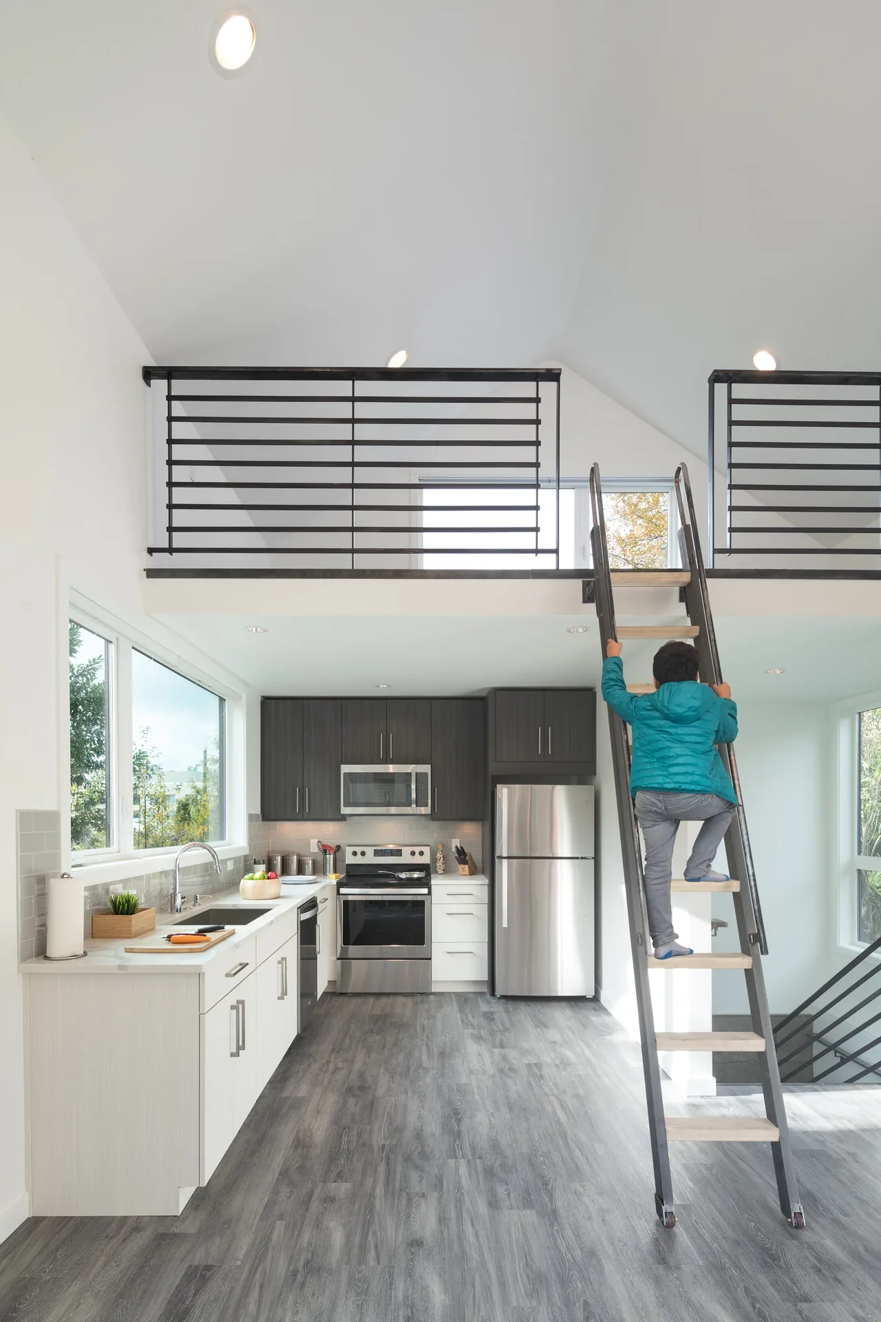 Modern kitchen with dark upper and light lower cabinets, stainless steel appliances, and a loft accessed by a black metal ladder.