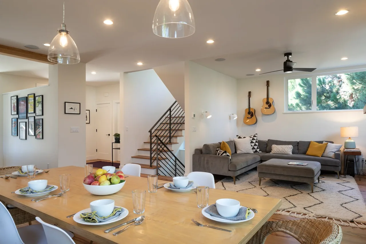 Living room and dining area with a wooden dining table, gray sectional sofa, and wall-mounted guitars under a wide window.