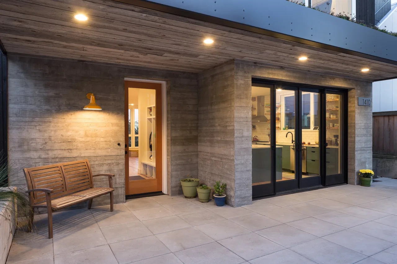 Backyard Residence patio features exposed concrete, a wooden bench, and black-framed glass doors to the kitchen interior.