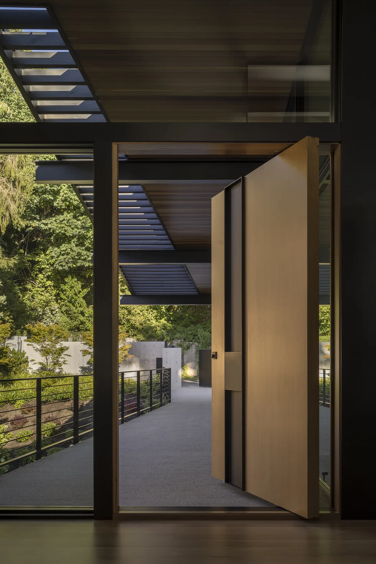 View through a pivot wooden entry door to an exterior balcony with metal railing and greenery beyond.