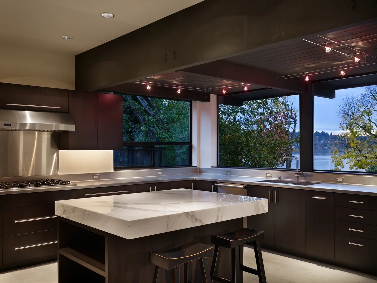 Lake Washington Residence kitchen by Baan Design features a marble island, dark cabinetry, stainless steel appliances, and large windows with lake views.