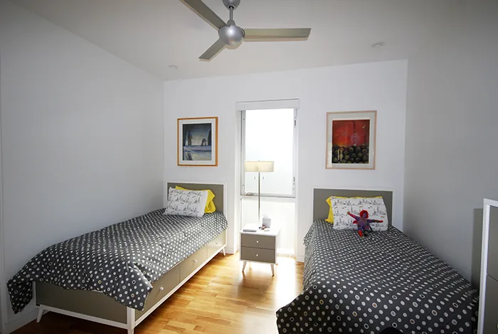 Twin beds flanking a window with a small nightstand and lamp in a bedroom with wood flooring and white walls.