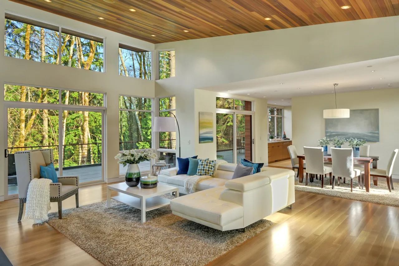 Open living and dining area with floor-to-ceiling windows overlooking a forest, white leather sectional sofa, and wood-paneled ceiling.