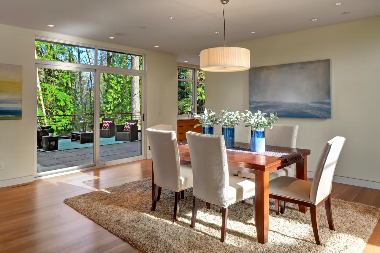 Dining room with wood table and beige chairs opens to an outdoor patio surrounded by forest views.