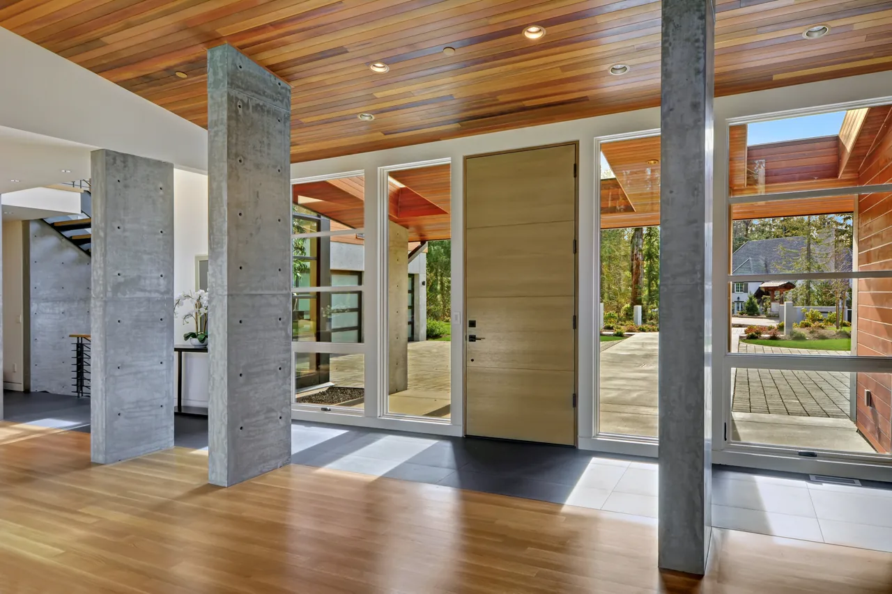 The entryway features concrete columns, a wood-paneled ceiling, and floor-to-ceiling glazing around a wooden front door.