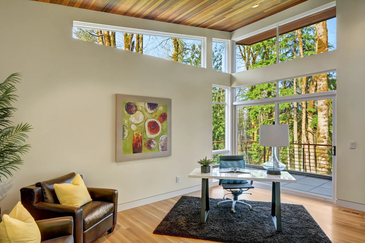 The Pavilion in the Woods home office features a wood paneled ceiling, large forest-facing windows, leather armchair, and a white desk with glass lamp.
