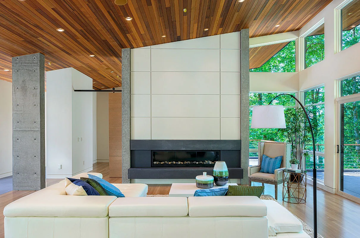 Living room interior with white sectional sofa centered before a modern fireplace, flanked by concrete columns and floor-to-ceiling windows.