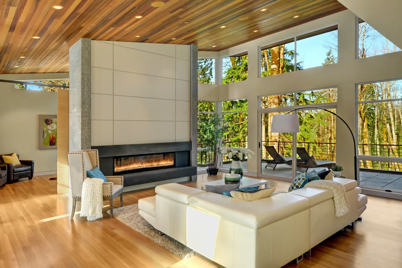 Open living room with floor-to-ceiling windows looking out to a wooded forest and a modern linear fireplace.