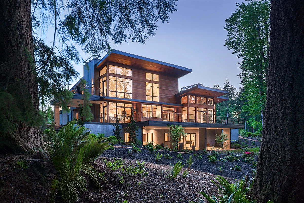 Modern multi-story house with extensive wood siding and floor-to-ceiling windows set among large trees at dusk.