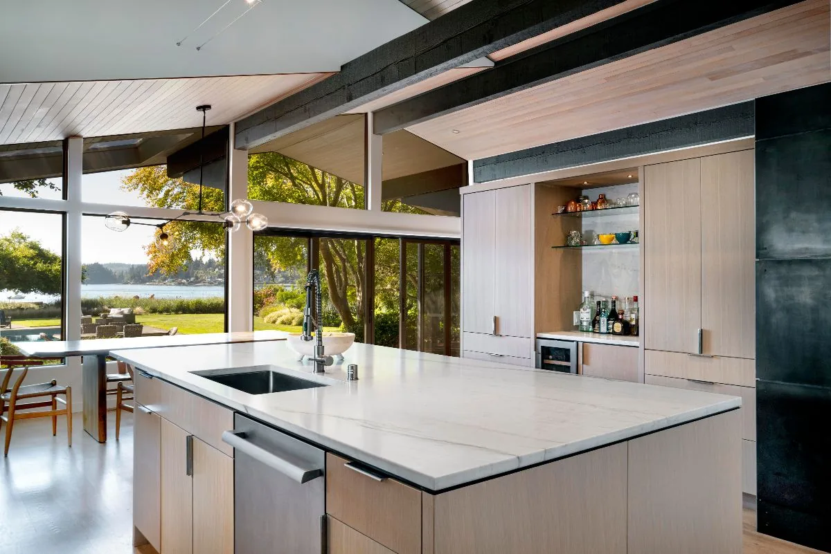 Mid-Century Beach House kitchen with marble island and wood-paneled ceiling overlooking a garden and waterfront.