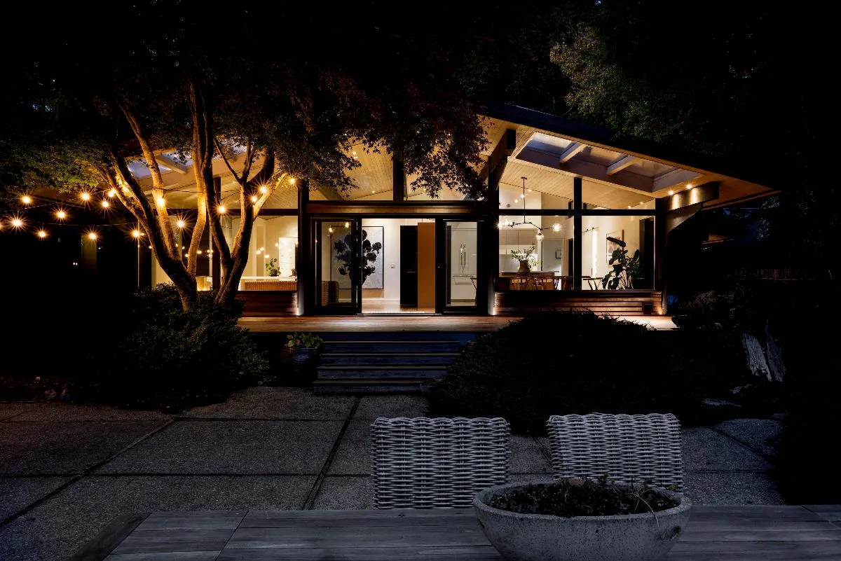 Mid-century modern beach house exterior lit at night with string lights and large glass windows showcasing interior spaces.