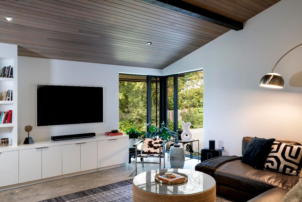 Living room in a Mid-Century Beach House features wood-paneled ceiling, corner windows, and a ribbed leather sofa with garden view.