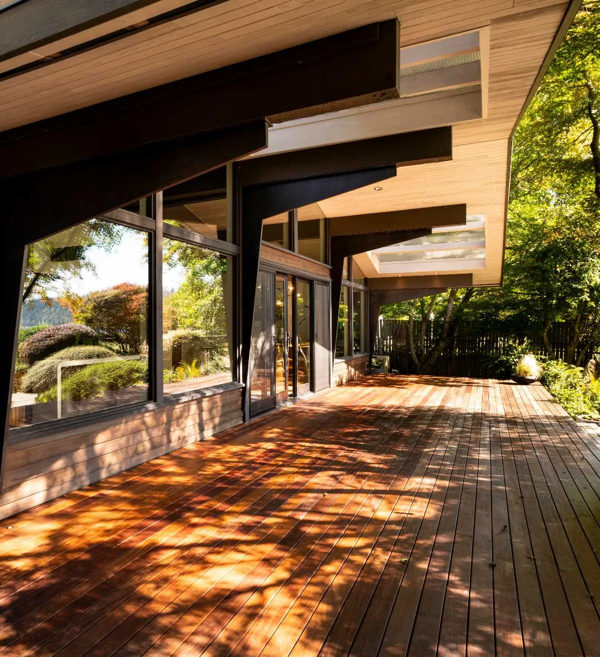 This outdoor living area features a broad wood deck with exposed beams extending from the ceiling and large glass windows showcasing garden views.