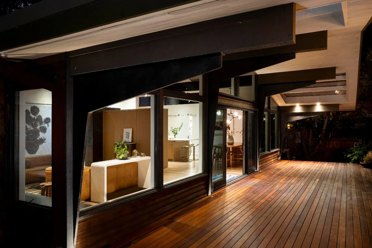 Mid-century modern house with a long wooden deck seen at night, featuring large glass windows and exposed beam ceilings.