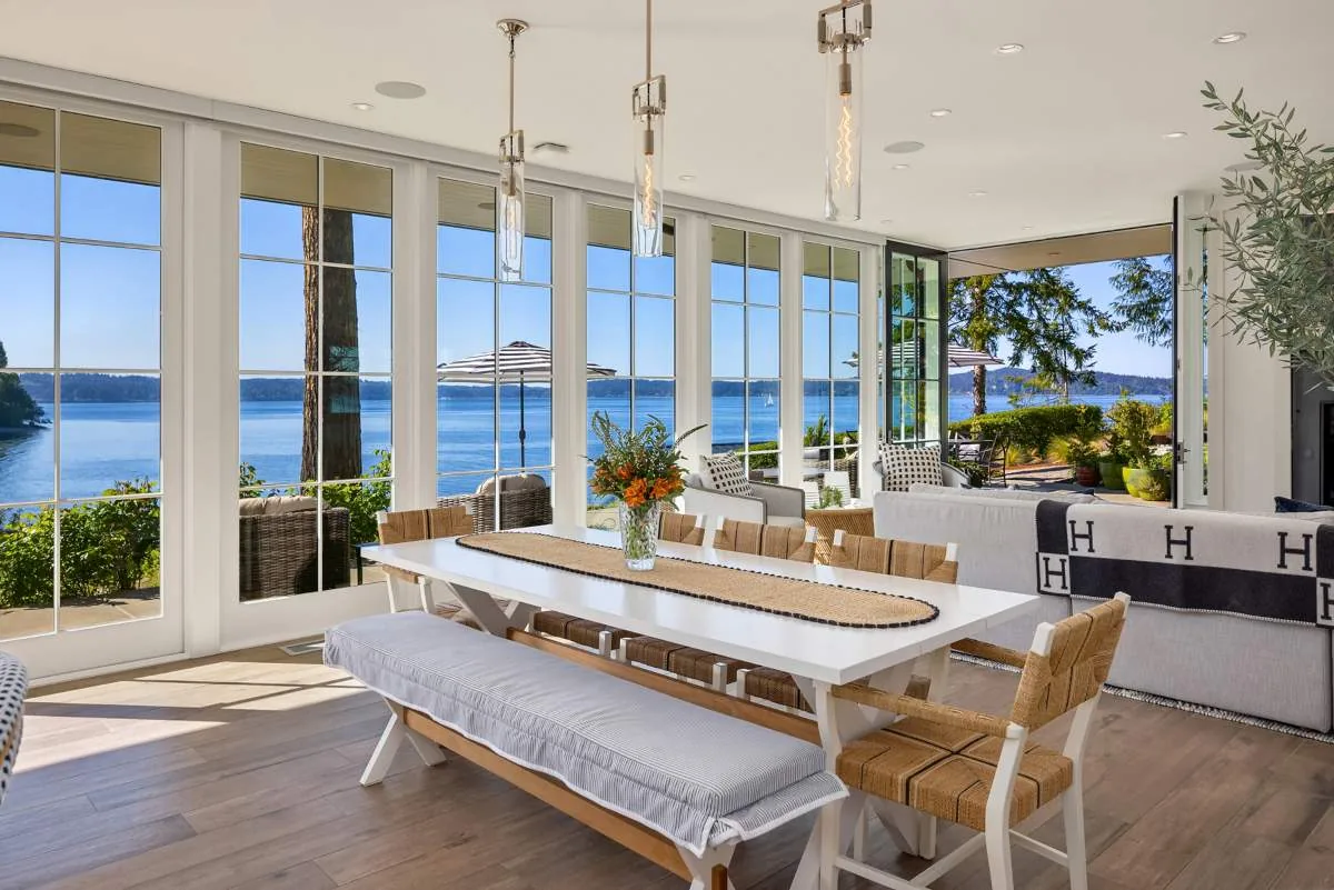 Dining room with floor-to-ceiling glazing overlooks water, featuring a white table, bench, and woven chairs.