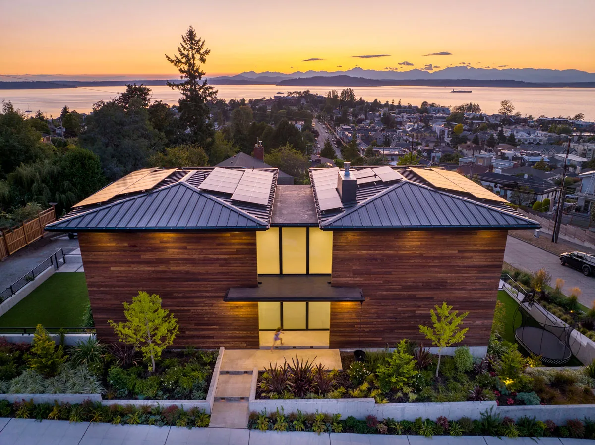 West Seattle House features wood siding and metal roofs with solar panels, situated on a sloped site with sunset lighting and a view of the water and mountains.