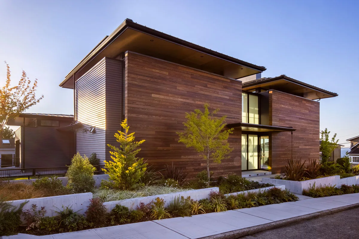 West Seattle House features horizontal wood siding and large glass windows with a flat roof, illuminated by sunset lighting.