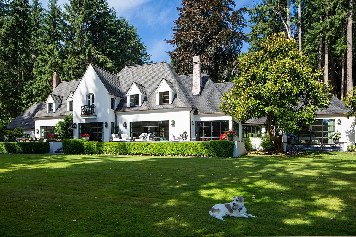 Manzanita Bay House features steep gable roofs and dormer windows, finished with white stucco and set in a lush natural landscape.