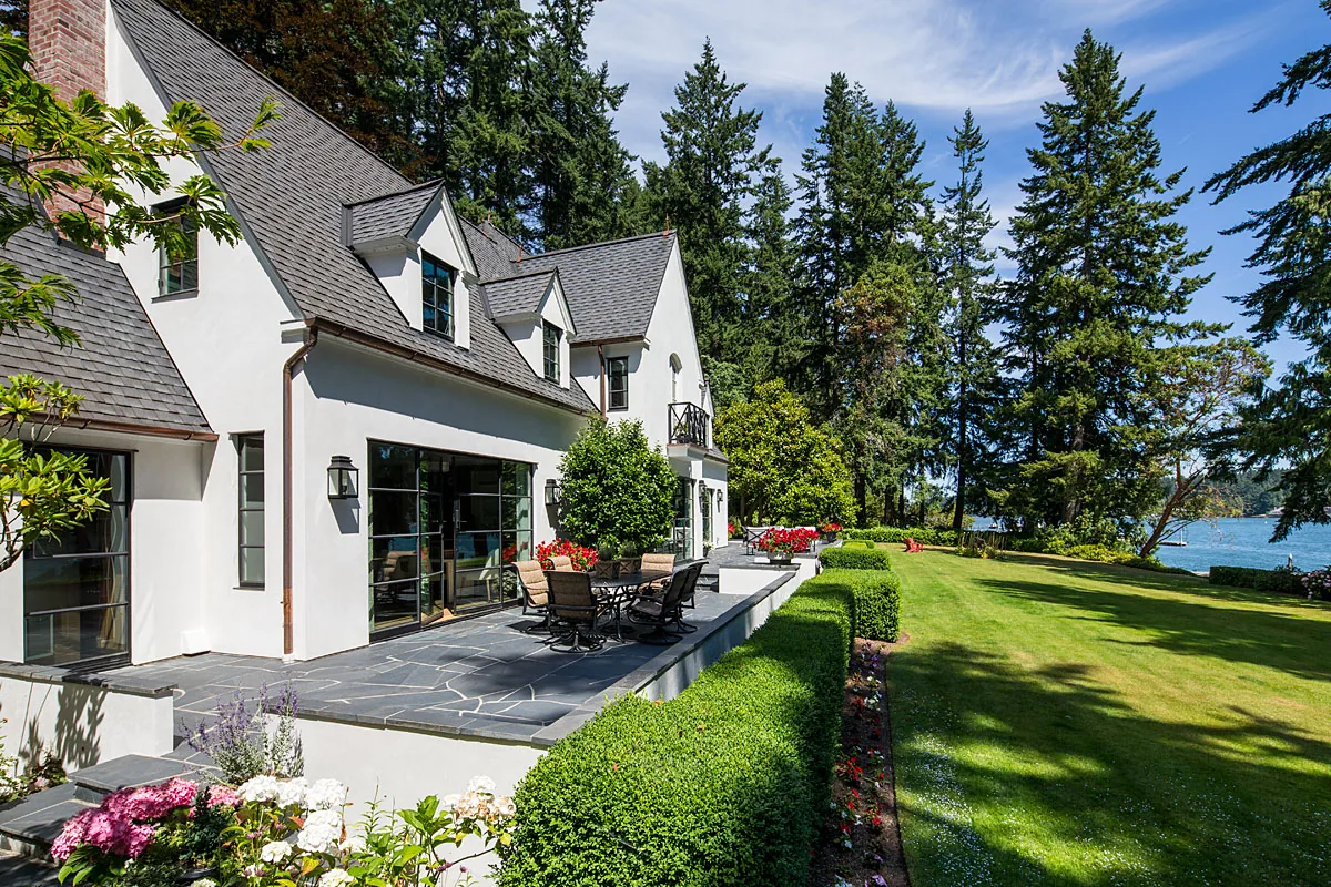 Manzanita Bay House features steep pitched roof with dormers, stone terrace with outdoor seating, and landscaped lawn leading to water view.
