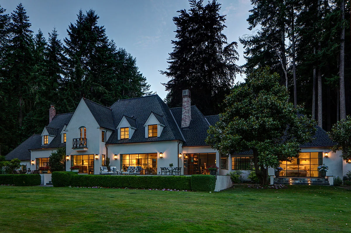 Evening view of Manzanita Bay House showing steep pitched roof, dormers, decorative chimney, small balcony, and landscaped lawn.