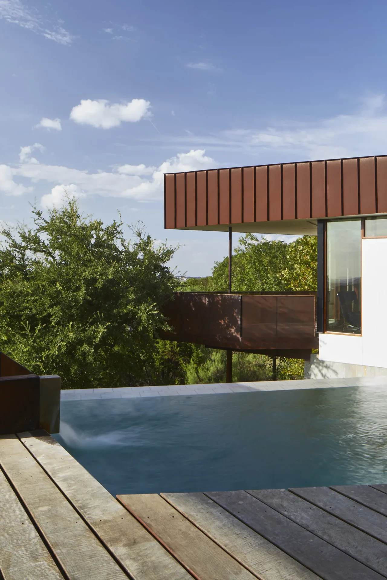 Infinity pool adjacent to a modern house with rust-colored metal siding and surrounded by trees