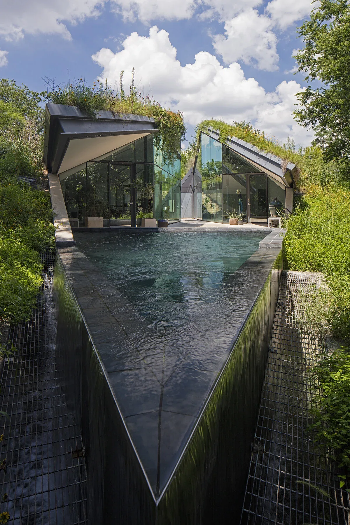 Bold Ruler House features a triangular reflective pool between two glass facades with sloped green roofs.
