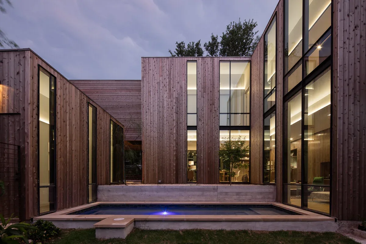 Evening view of a modern courtyard house with vertical wood siding and tall windows surrounding a rectangular pool.
