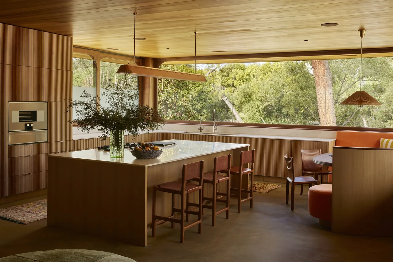 Claremont residence kitchen features a large island, curved banquette seating, and rounded window frames with views of lush landscaping.