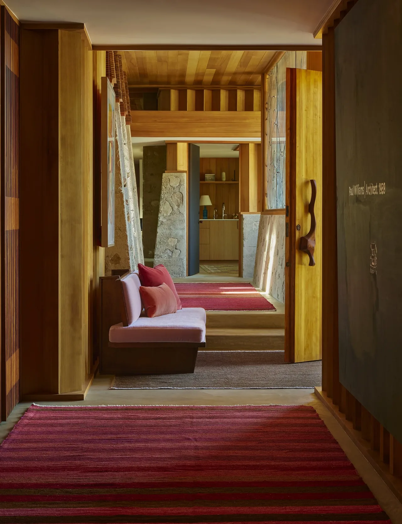 A hallway with wood paneling, red striped rugs, and built-in bench seating leads to a kitchen in the Claremont residence.
