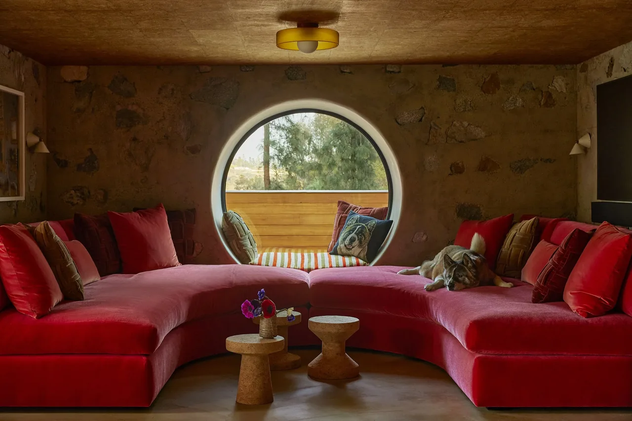 Living room of Claremont residence features a curved red sofa and a distinctive round window with wood paneling outside.