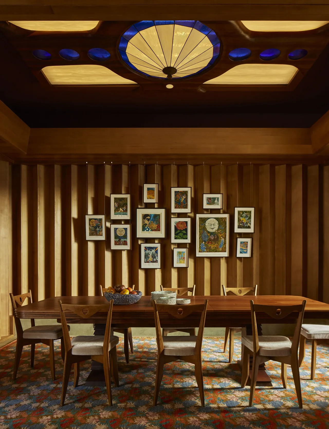 Dining room features wood-paneled slatted wall with framed art, midcentury style wooden chairs, and a patterned rug beneath a stained glass ceiling light.