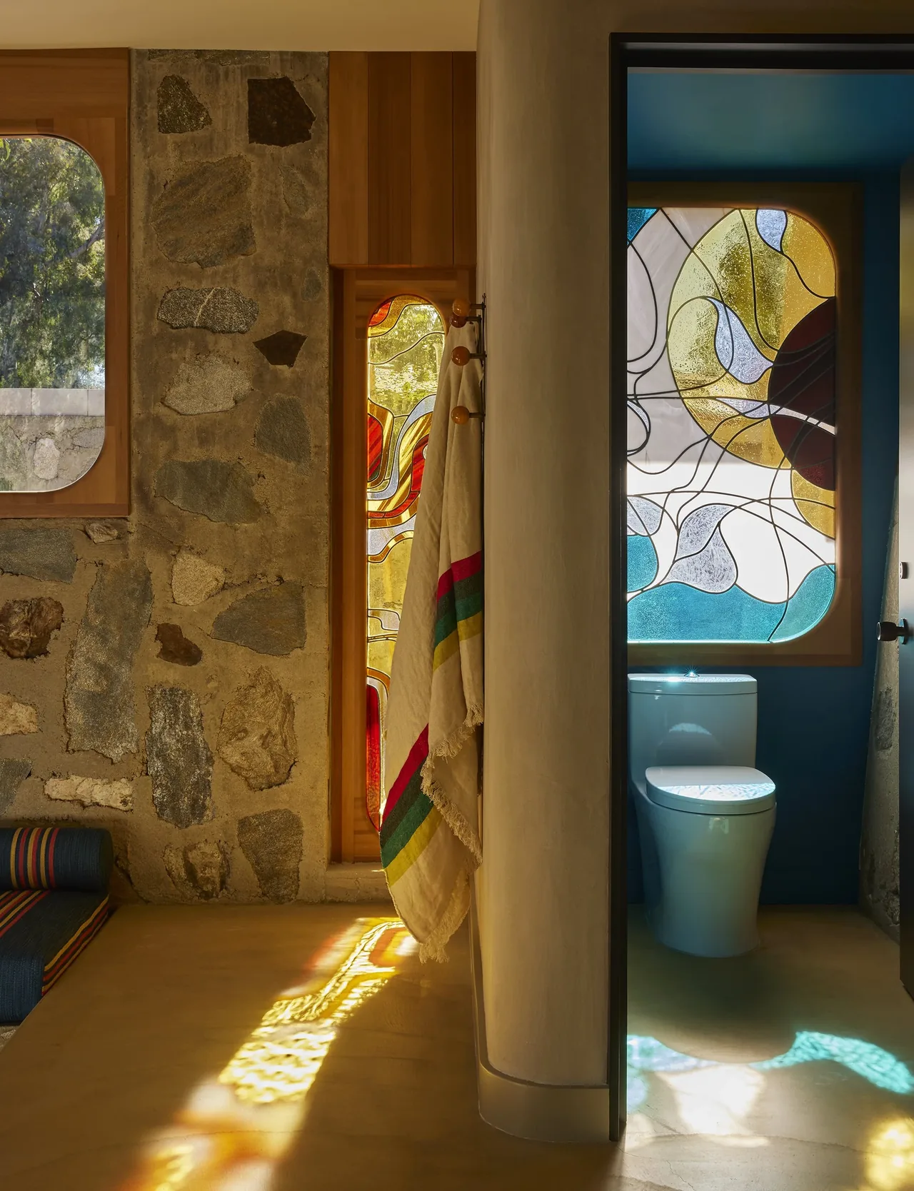 Room with stone and wood walls and a bathroom with stained glass window and toilet, showing light patterns on the floor.