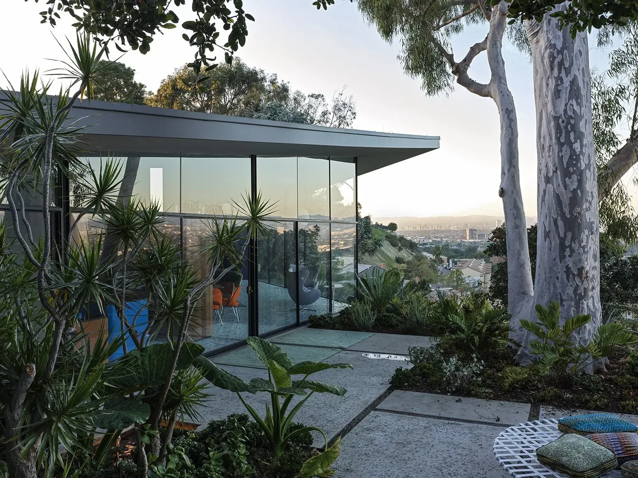 Exterior view of a modern home with extensive glass walls and a cantilevered flat roof overlooking a hillside at sunset.