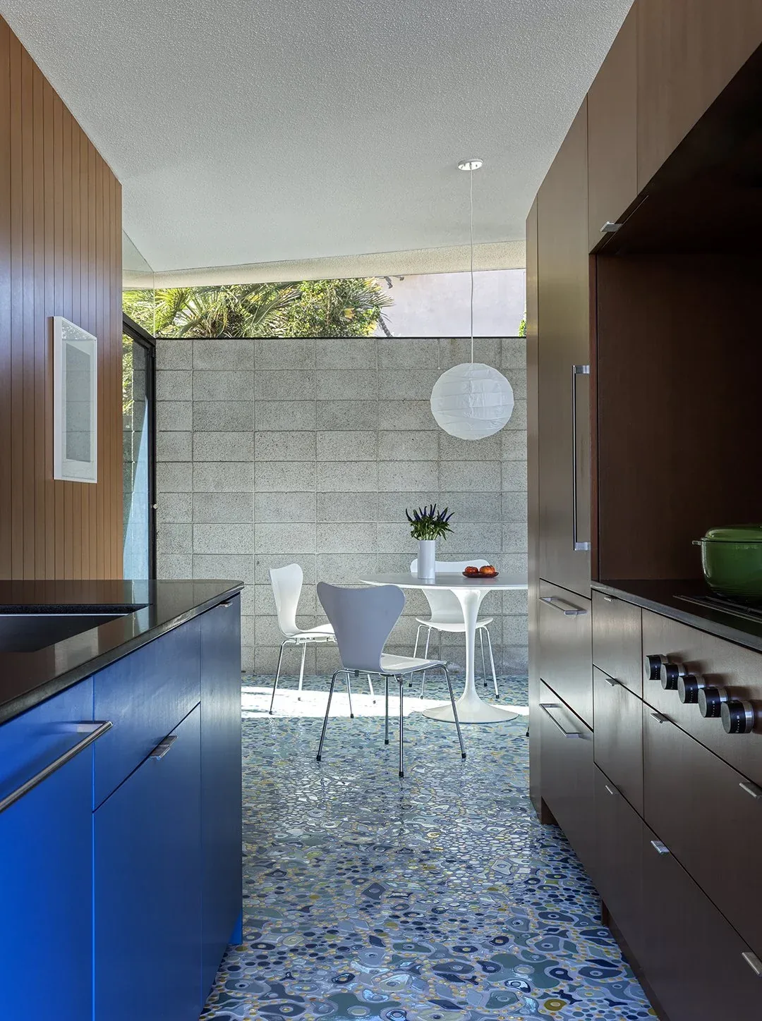 This kitchen features blue lower cabinets, wood paneling, terrazzo flooring, and a small dining area with white pedestal table and chairs.